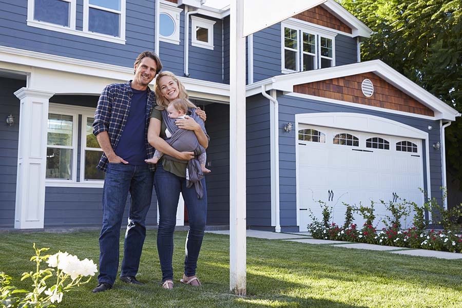 Personal Insurance - Young Smiling Parents Holding a Baby Standing in Front of Their New Two Story Home